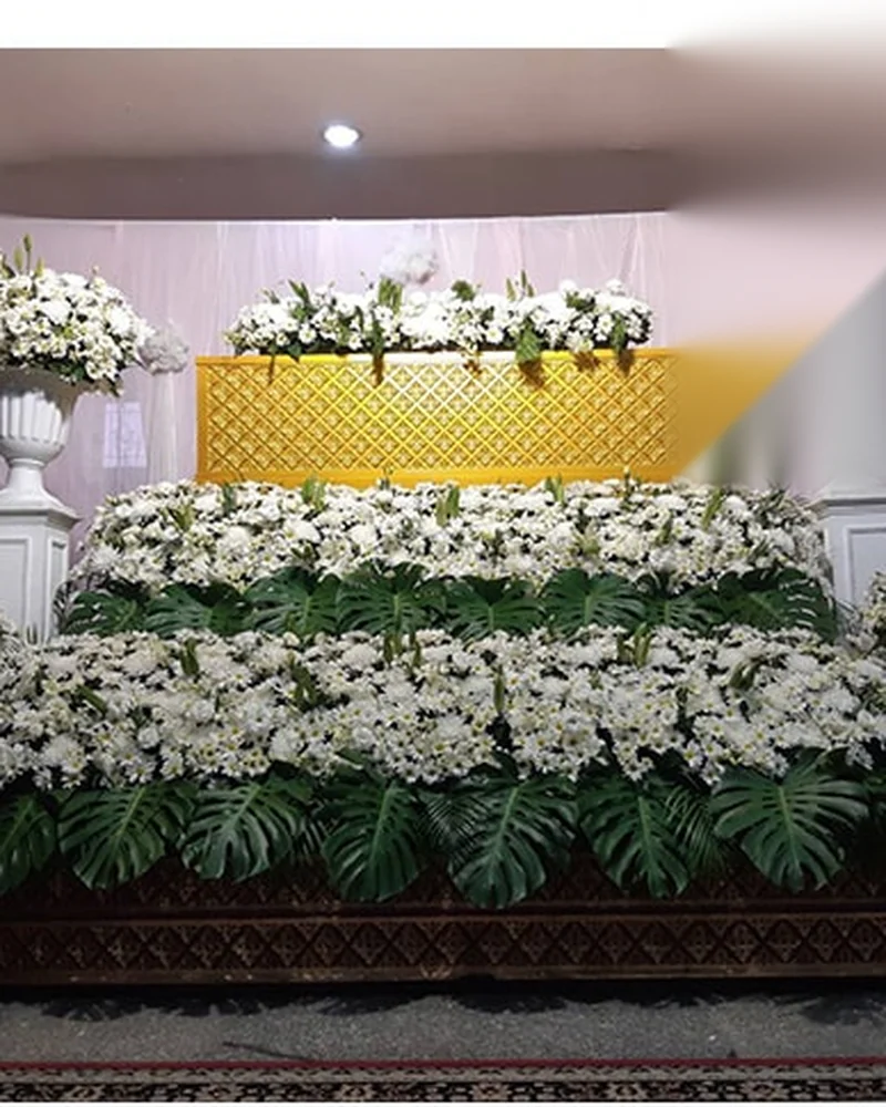 ดอกไม้สีขาวจัดเรียงเป็นชั้นประดับใบมอนสเตอร่าเขียวรอบขอบในศาลาสวดพระอภิธรรม