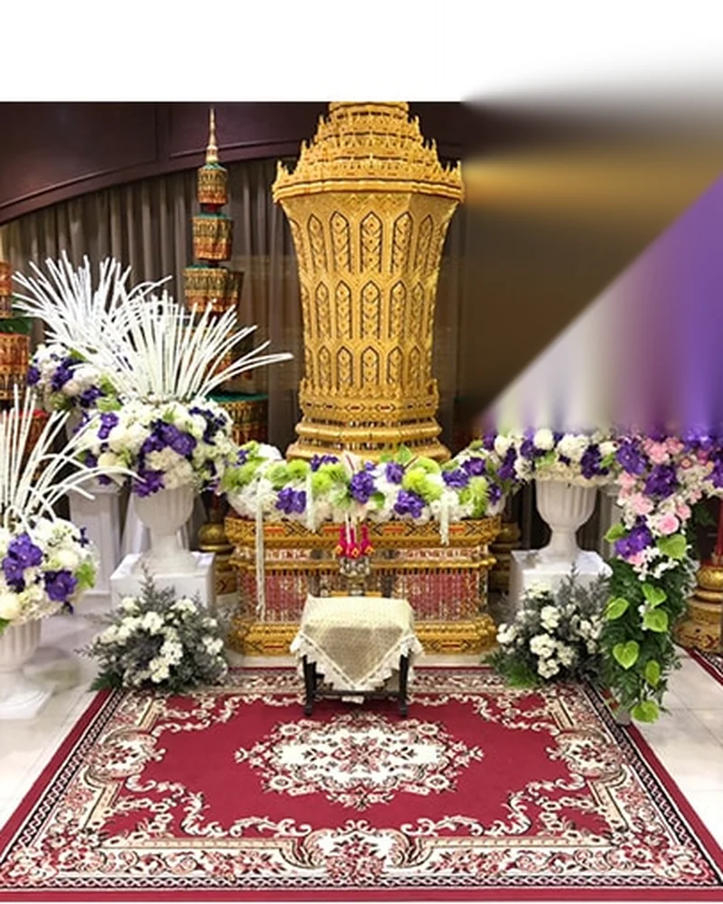 ดอกไม้สีม่วงขาวประดับรอบแท่นทองลวดลายไทยประเพณีบนพรมสีแดง