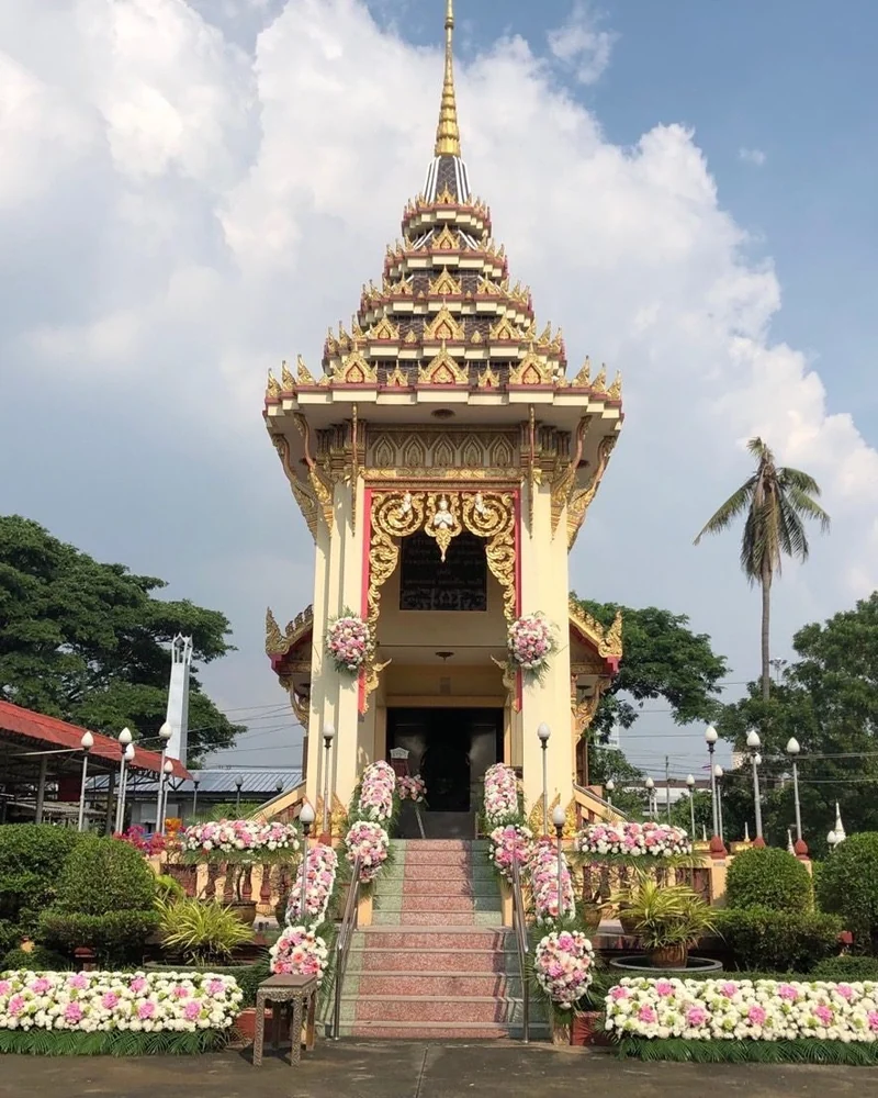 เมรุสีทองประดับดอกไม้สีชมพูขาวสองข้างบันไดทางขึ้น ท้องฟ้าสดใสมีต้นมะพร้าว